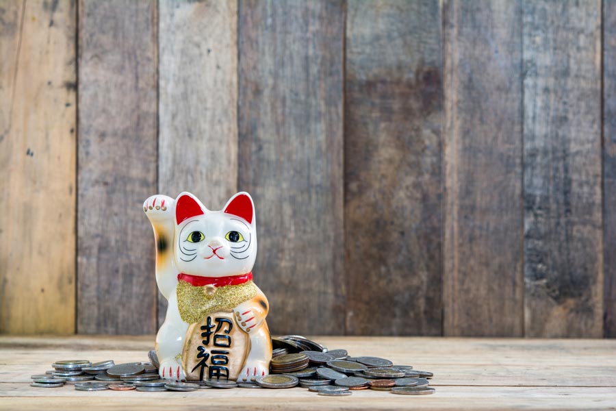 White ceramic cat statuette with one paw raised. The cat is on a wooden table and is surrounded by gray coins.