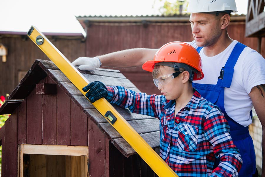 A man and a child wearing carpenter's work clothes are building a doghouse